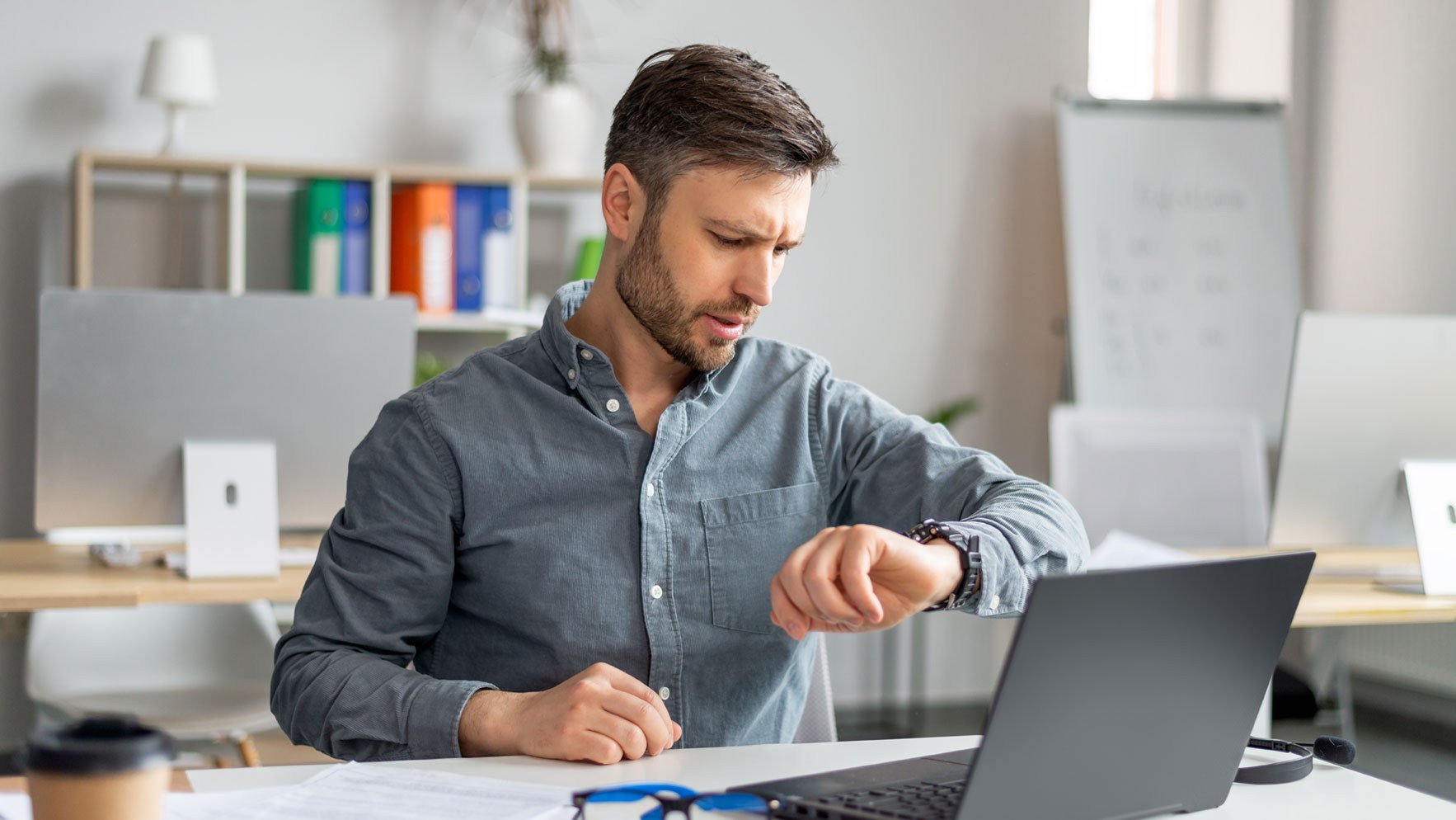 Ein Arbeitnehmer sitzt am Schreibtisch vor seinem Laptop und blickt auf seine Armbanduhr.
