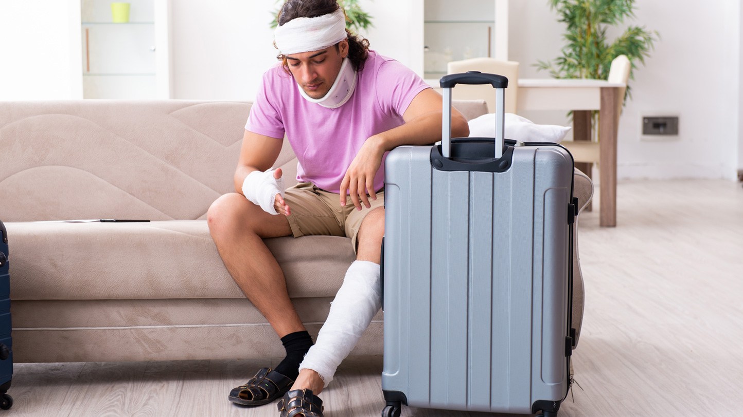 Ein Mann mit Verbänden an Arm, Fuß und Kopf sitzt neben einem Reisekoffer.