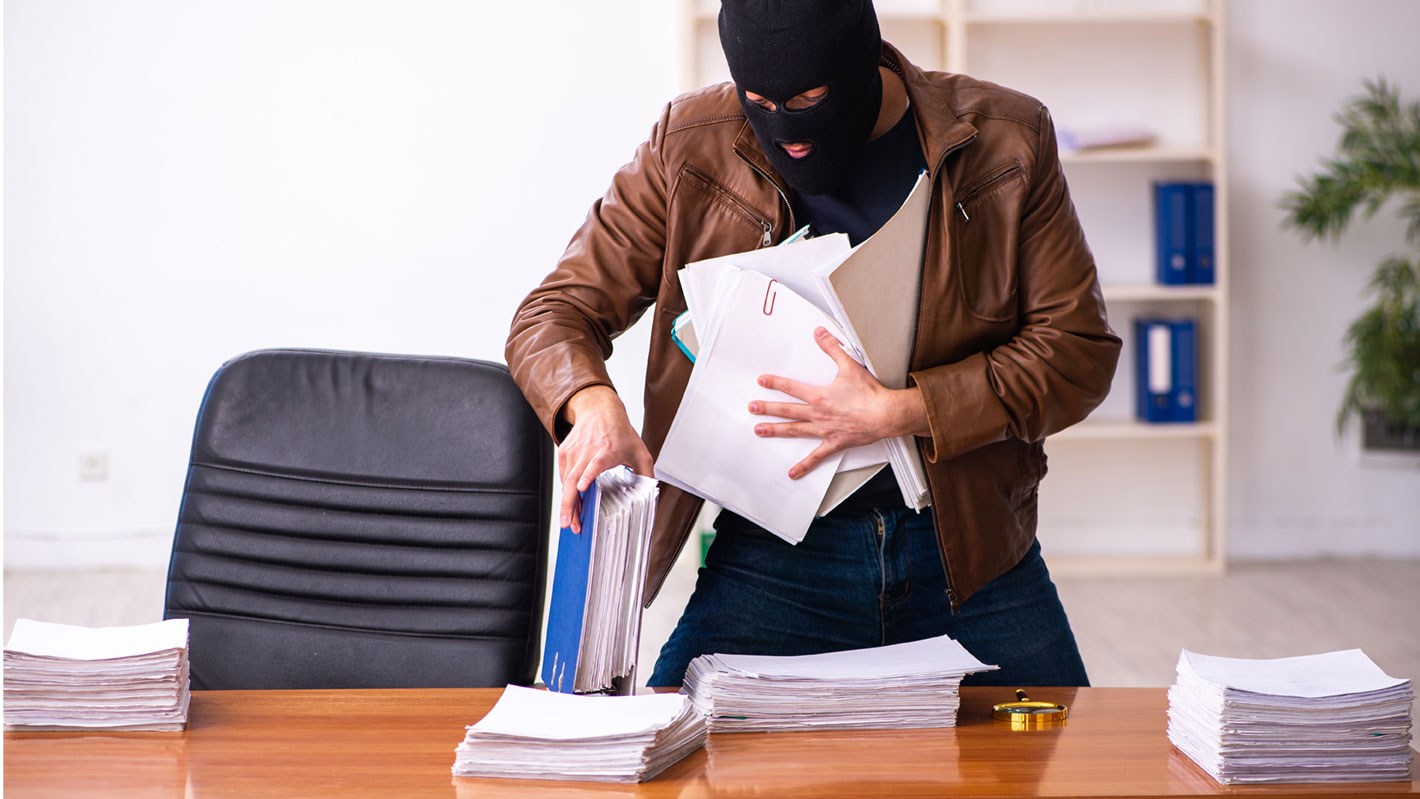 Ein Mann mit Maske stiehlt Bürounterlagen von einem Schreibtisch.