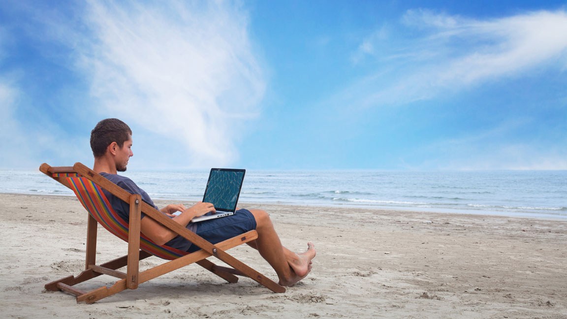 Ein Mann sitzt in einem Liegestuhl am Strand. Er hat einen Laptop auf dem Schoß.