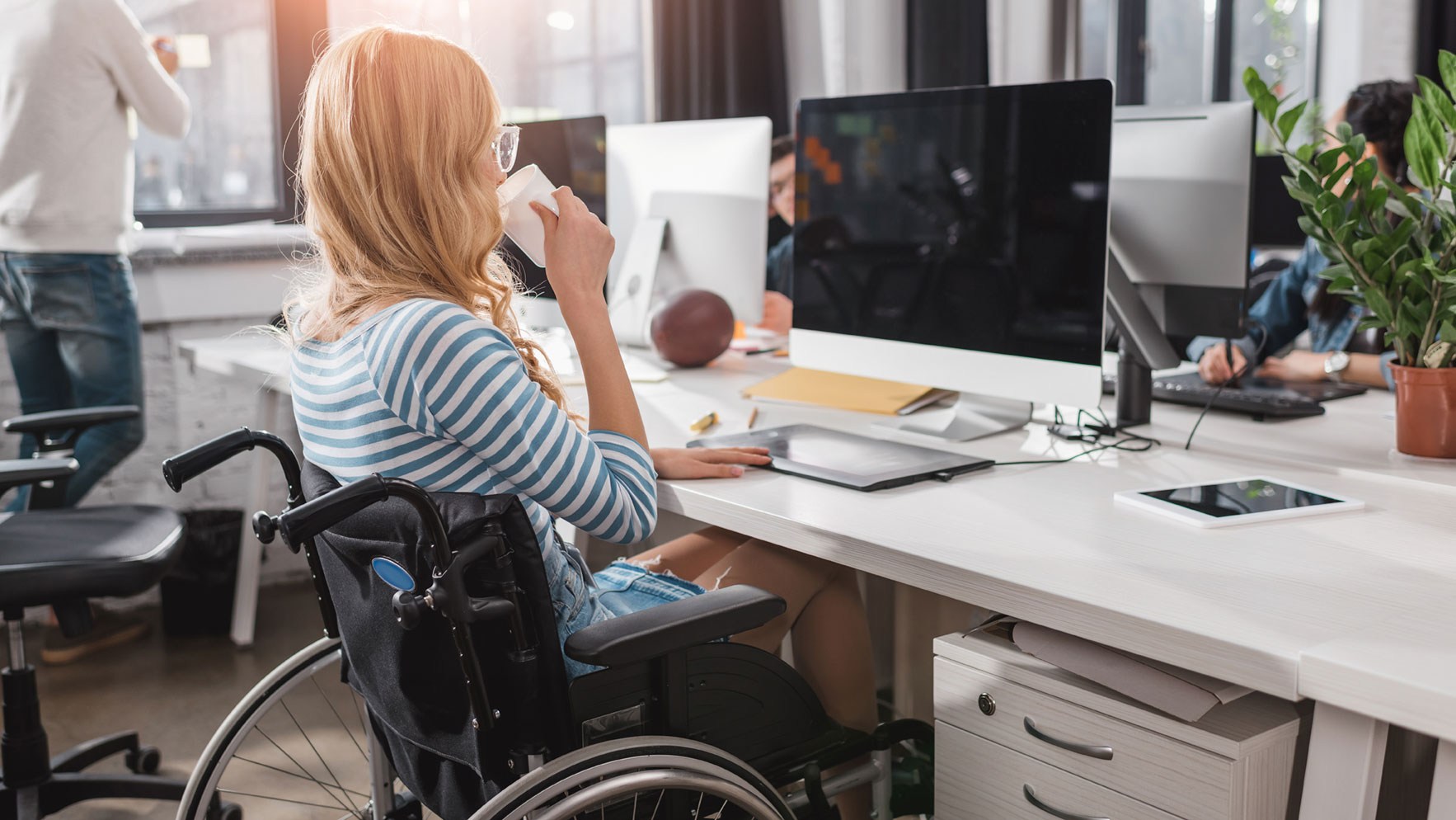 Eine Arbeitnehmerin im Rollstuhl sitzt im Büro und arbeitet am Computer.