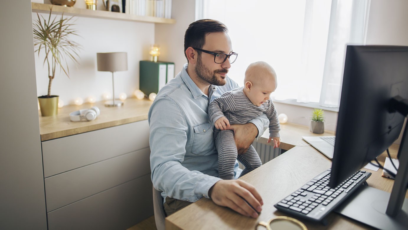 Ein Mann sitzt am Schreibtisch und arbeitet. Er hält ein Kleinkind auf dem Arm.
