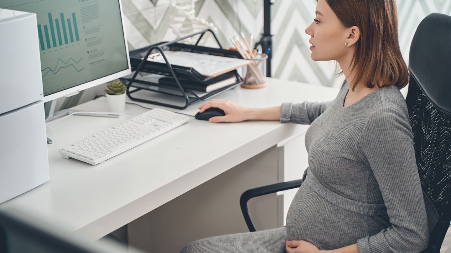 Eine schwangere Arbeitnehmerin sitzt im Büro und arbeitet an ihrem Computer.