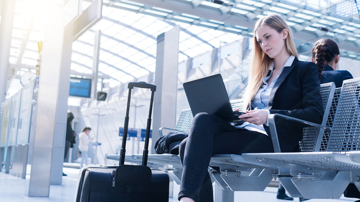 Eine Arbeitnehmerin sitzt mit Laptop und Koffer am Bahnhof.