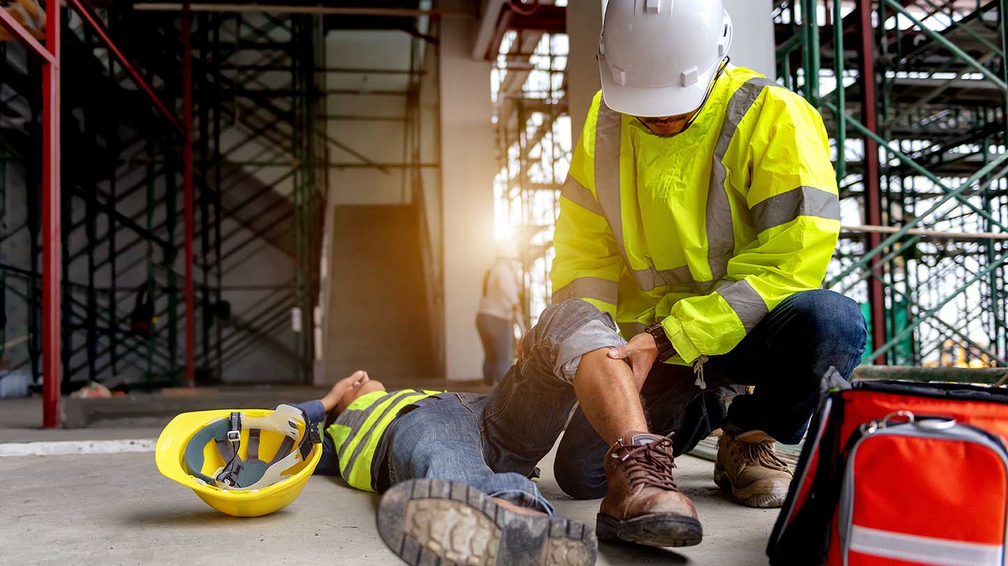 Ein Arbeitnehmer hatte einen Arbeitsunfall und liegt am Boden. Ein Kollege leistet Erste Hilfe.