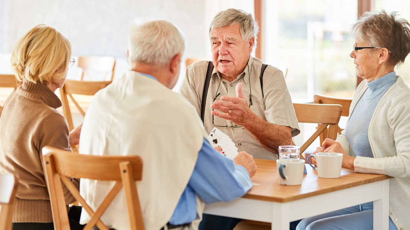 Vier Senioren sitzen an einem Tisch. Sie trinken Kaffee und spielen Karten