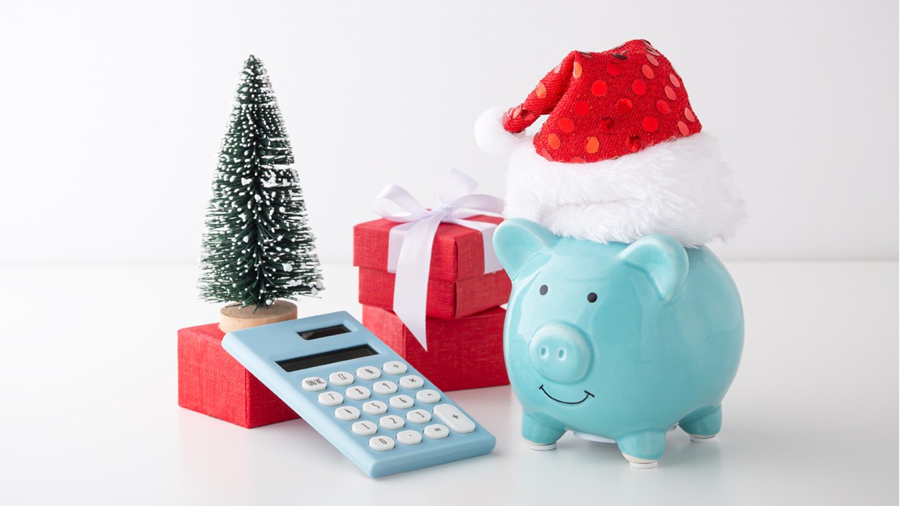 Ein Sparschwein mit Weihnachtsmütze, das neben einem Taschenrechner und Weihnachtsgeschenken steht.